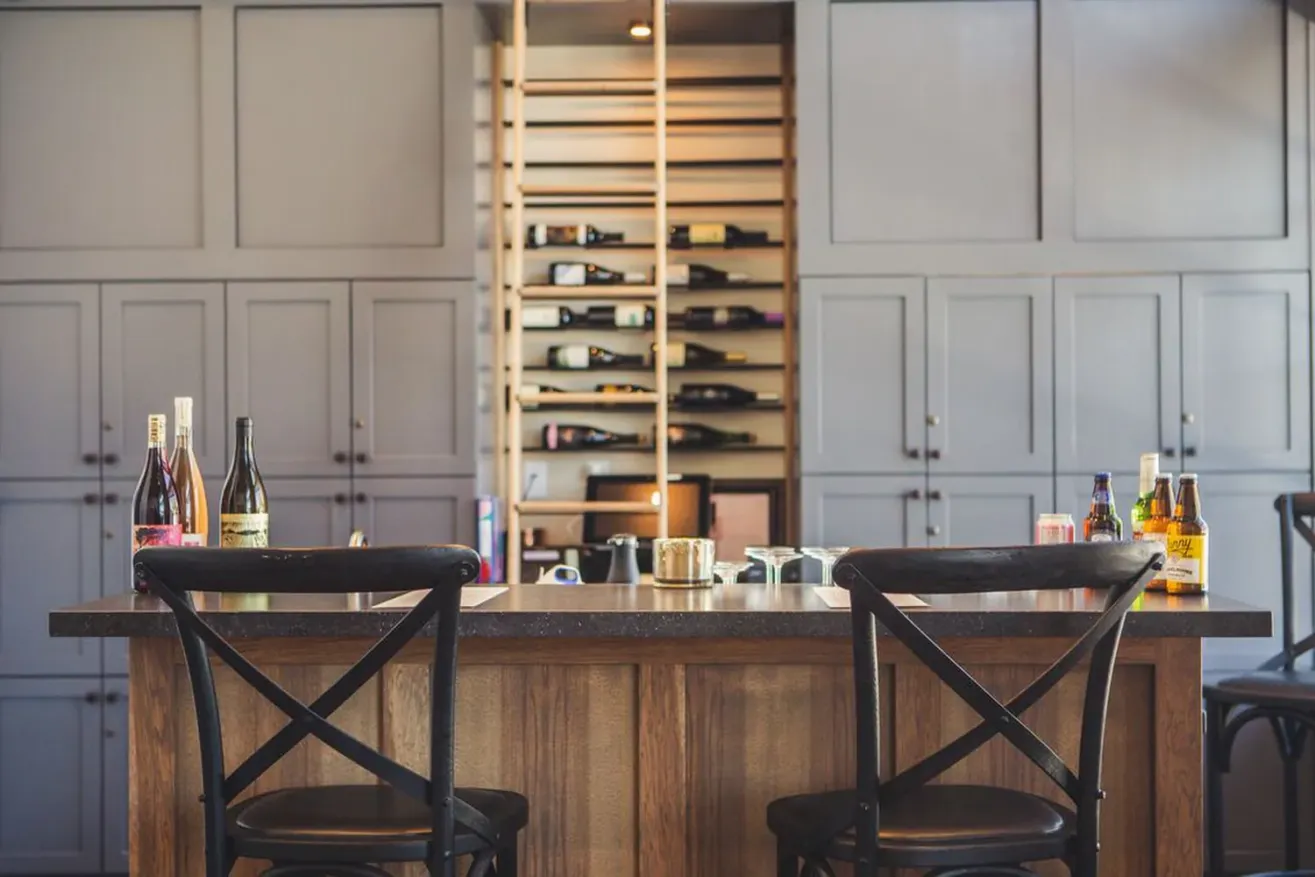 A modern bar area at a boutique hotel in Paso Robles, featuring two black chairs at a wooden counter, bottles of wine, liquor, and mixers on the countertop, with shelves and wine bottles displayed in the background.