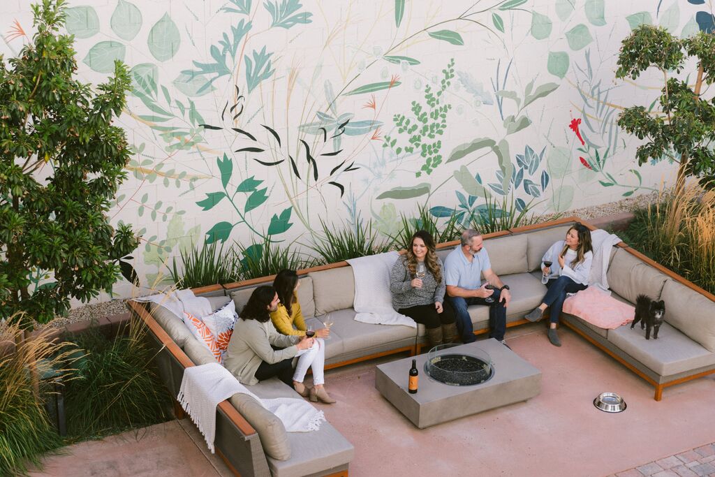 Five people sit and chat on outdoor couches around a fire pit in a cozy patio at The Piccolo at Paso Robles Inn, decorated with a large botanical mural and surrounded by greenery; a small black dog stands nearby.
