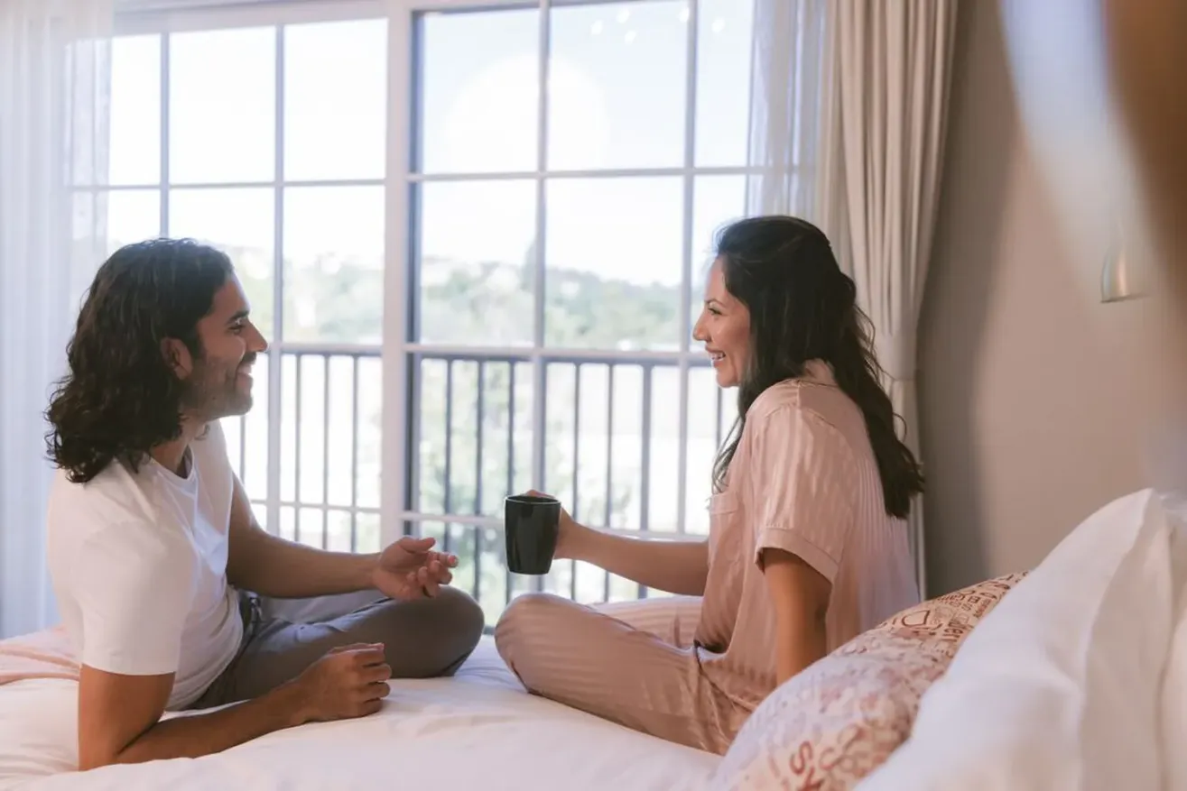 A man and woman sit on a bed by a large window at The Piccolo at Paso Robles Inn, smiling and talking. The woman holds a black mug, and both appear relaxed and happy in the bright, cozy bedroom.