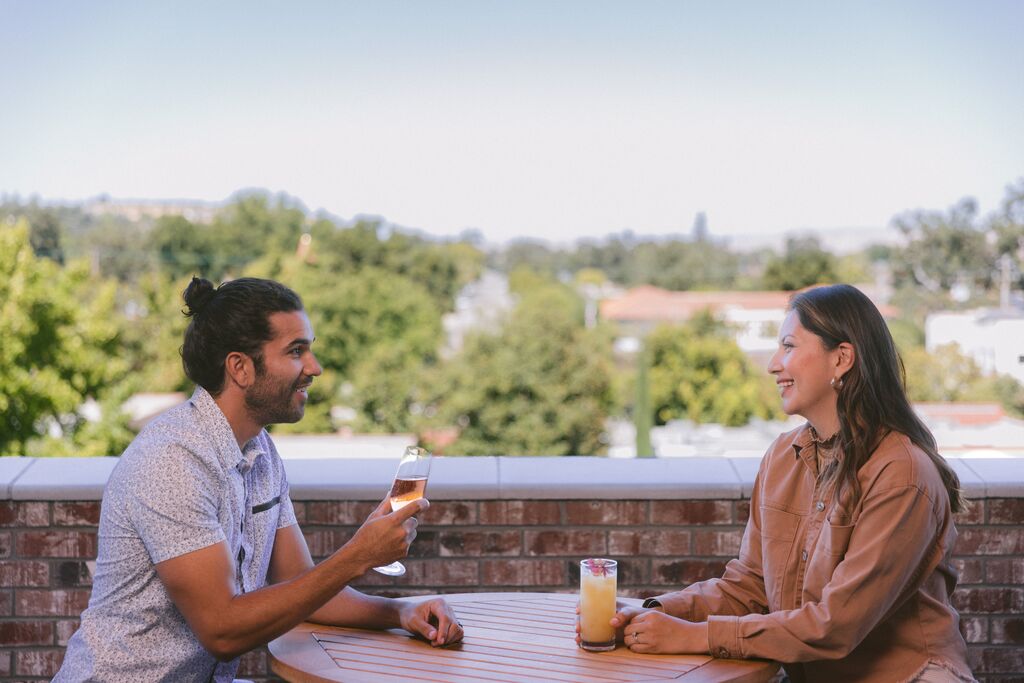 Two people sit at a round outdoor table at The Piccolo at Paso Robles Inn, smiling and talking with drinks in hand. Trees and buildings are visible in the background under a clear sky.