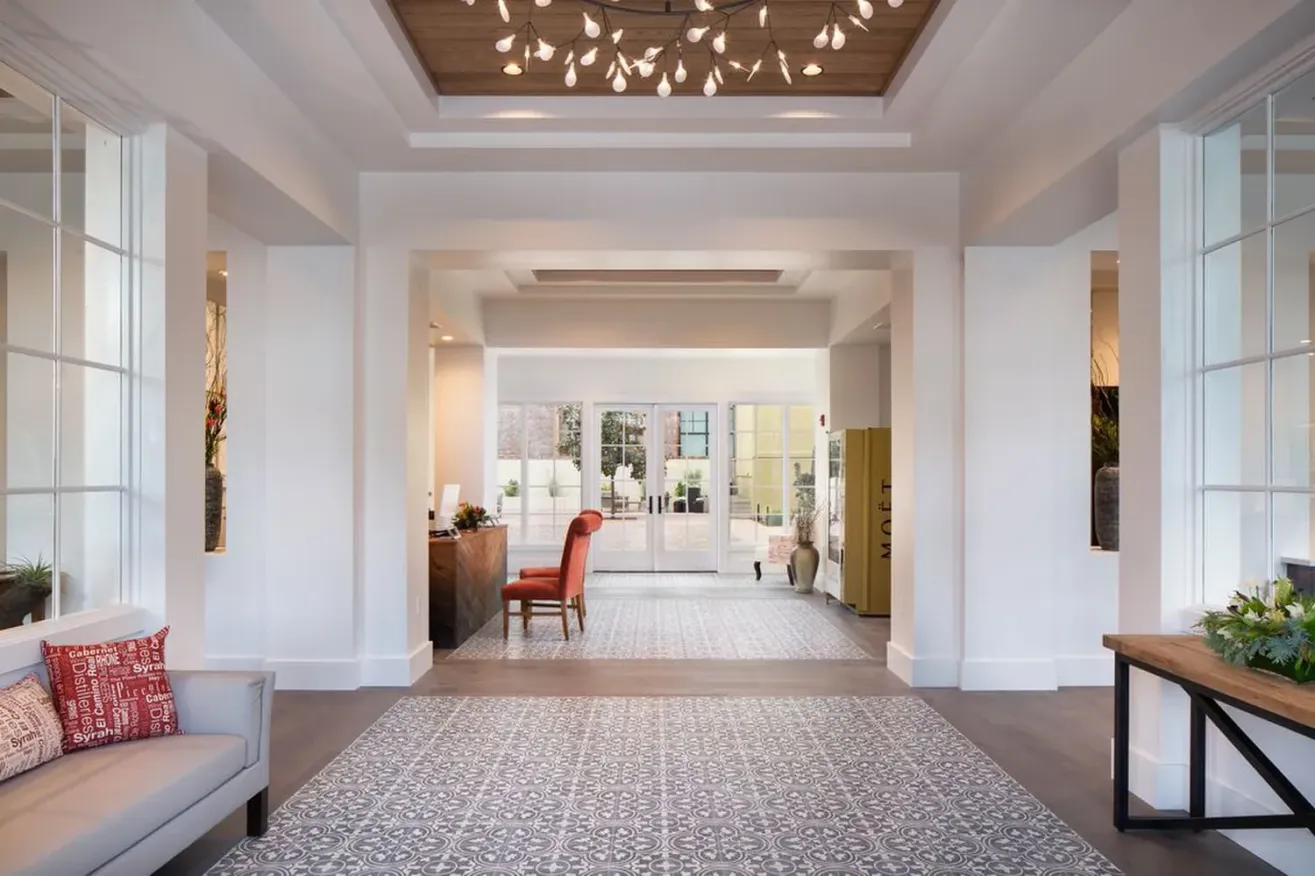 A bright, modern lobby in a boutique hotel in Paso Robles features a patterned tile floor, white walls, large windows, and wood ceiling accents with hanging lights. A red chair sits at a desk by glass doors opening to a patio with greenery.