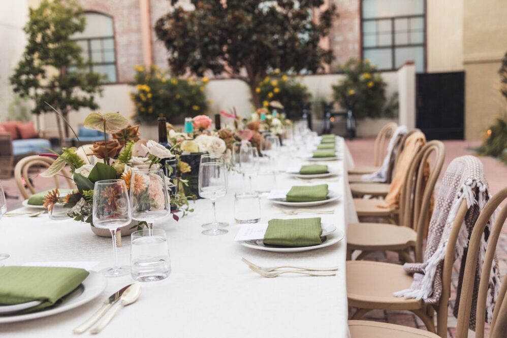 A long outdoor dining table with a white tablecloth is set with green napkins, glassware, and floral centerpieces, surrounded by wooden chairs in a courtyard at The Piccolo at Paso Robles Inn, nestled among trees and blooming plants.