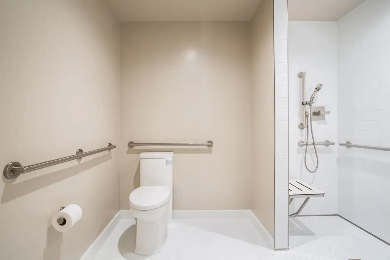 An accessible bathroom with beige walls, featuring a toilet with grab bars, a toilet paper holder, and a roll-in shower with a fold-down seat, grab bars, and a handheld showerhead.