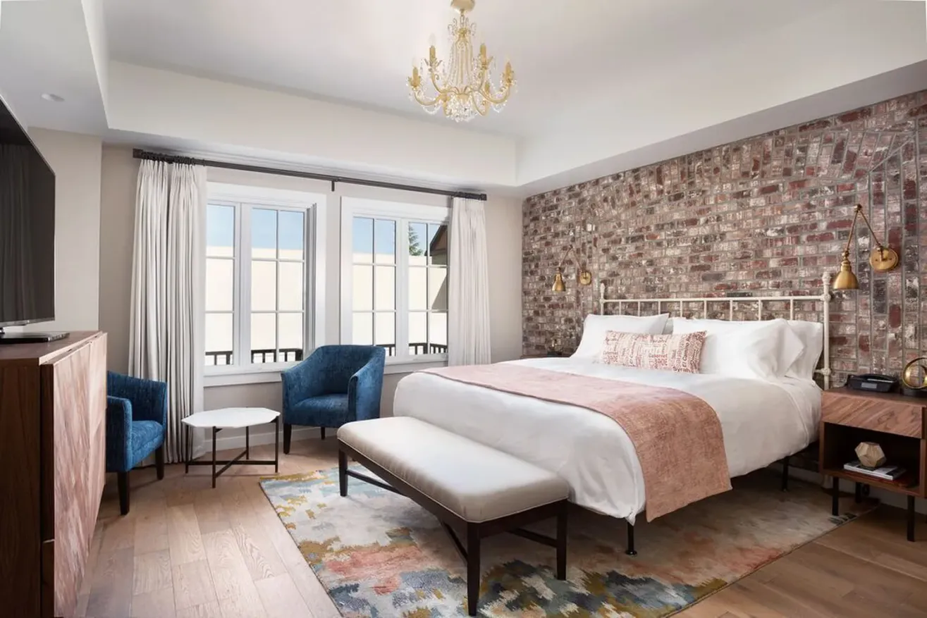 A stylish bedroom with a brick accent wall, white bed with pink throw, blue chairs, a bench, rug, chandelier, and large window with white curtains letting in natural light.