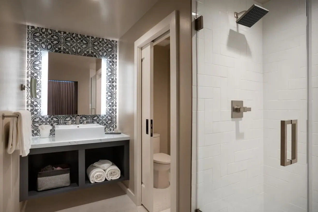Modern bathroom with a patterned tile accent wall, illuminated mirror above a sink with storage shelves, visible towels, and a glass-enclosed shower with a square showerhead.