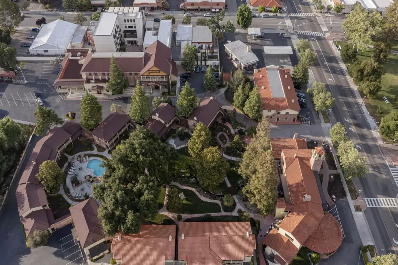 Aerial view of a hotel complex with red-roofed buildings surrounding a garden with a pool, set near a city street and surrounded by trees and parked cars.