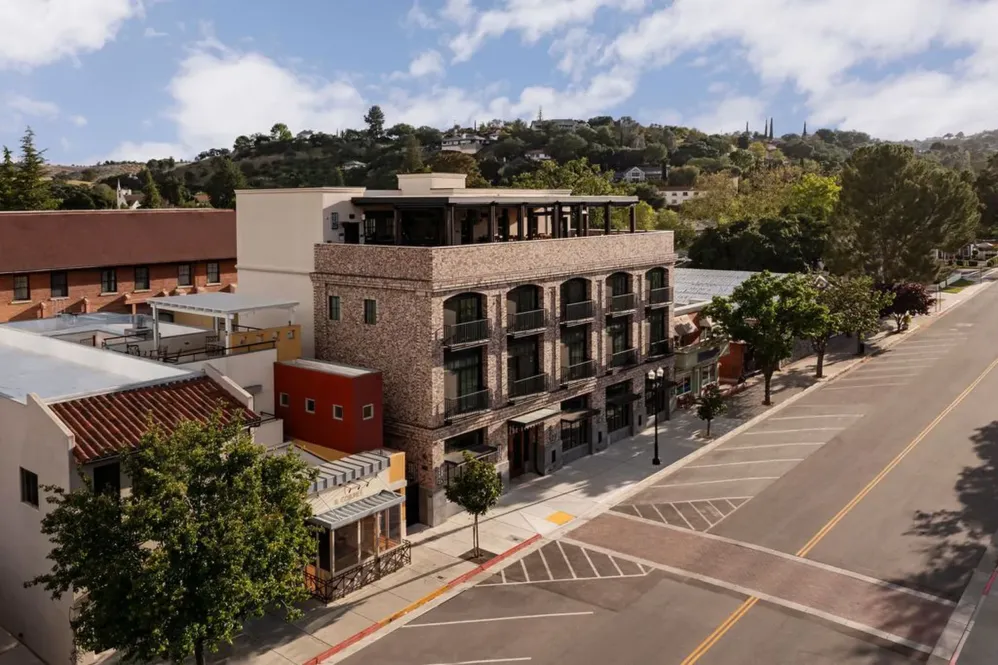 A three-story brick building with balconies lines a quiet, tree-lined street on a sunny day; hills with houses and greenery are visible in the background under a partly cloudy sky—perfect for guests seeking the Paso Government Rate.