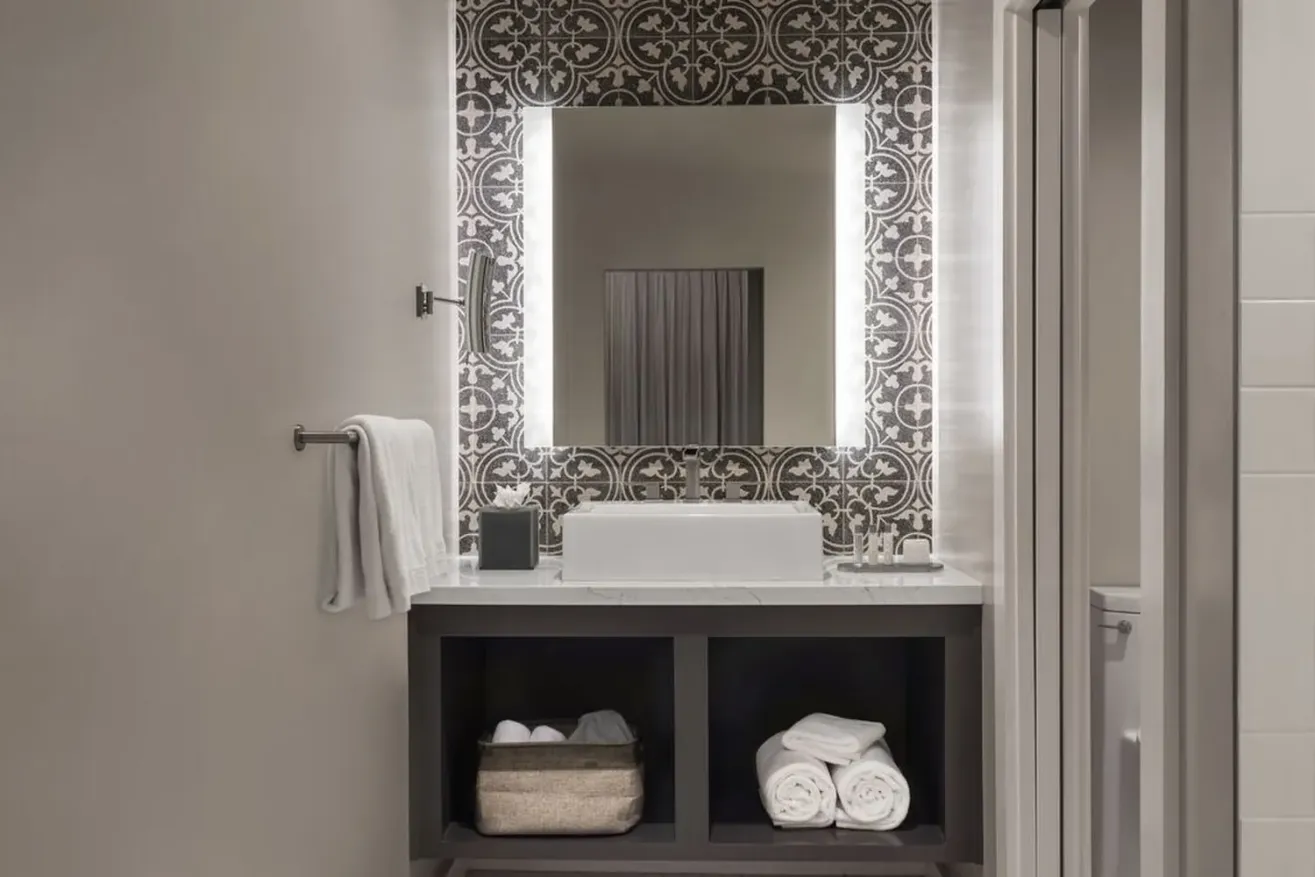 Modern bathroom with patterned tile wall, large illuminated mirror, white sink on a dark vanity, rolled towels and a basket stored on open shelves below. A hand towel hangs on a nearby metal bar.