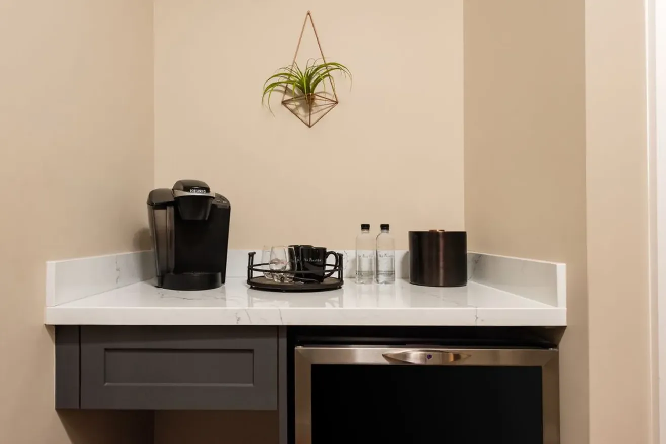 A small coffee station with a Keurig coffee maker, two water bottles, cups on a tray, and an ice bucket on a white countertop. A plant in a geometric wall holder hangs above.