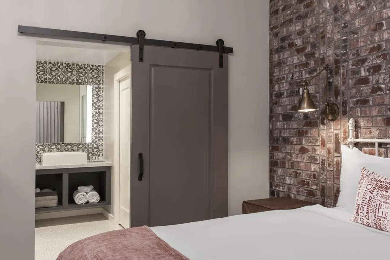 A cozy bedroom with a brick accent wall, a bed with white linens and decorative pillow, a wooden nightstand, and a sliding barn door leading to a bathroom with a vanity and patterned tiles.