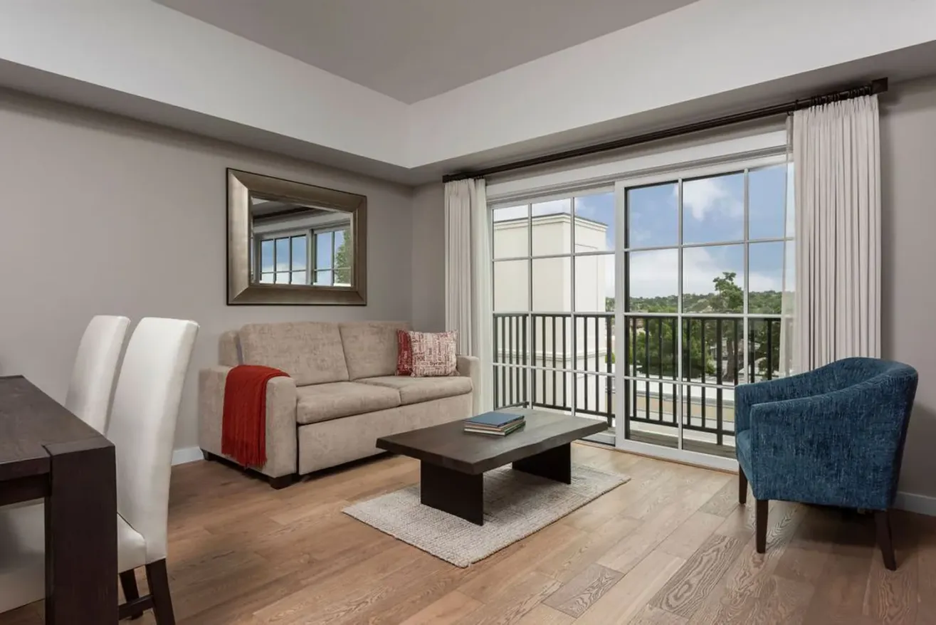 Modern living room in suites in Paso Robles features a beige sofa, red throw, blue accent chair, coffee table, and dining area. Large window lets in natural light, revealing a balcony and greenery. Neutral palette and minimalist decor create a relaxing vibe.