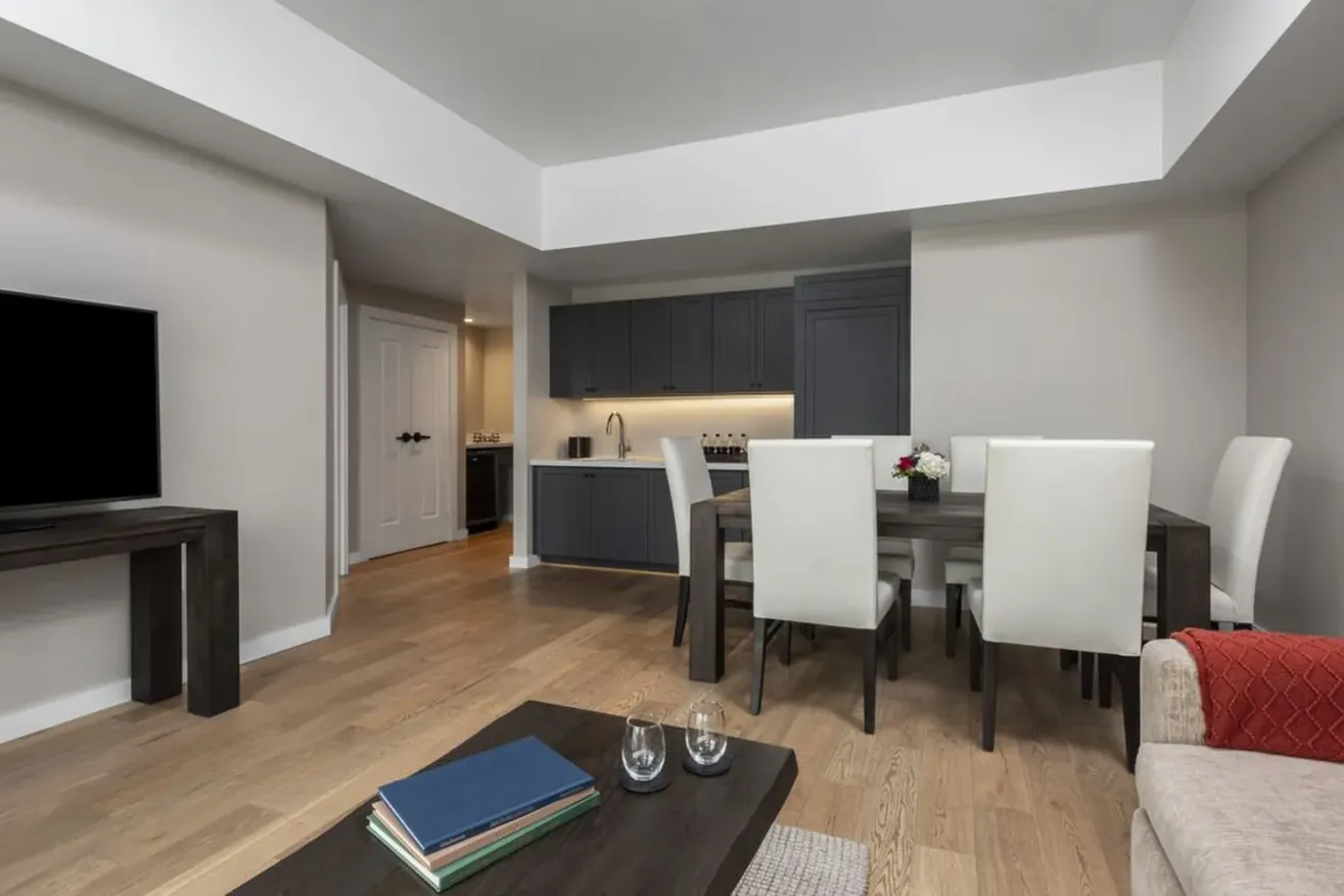 Modern open-plan living and dining area with light wood flooring, a dark wood dining table with six white chairs, a kitchenette with gray cabinets, and a beige sofa with a red pillow.