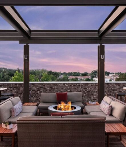 Outdoor rooftop lounge at The Piccolo at Paso Robles Inn features modern furniture around a circular fire pit beneath a pergola, overlooking the cityscape and treetops at sunset beneath a vibrant purple and pink sky.