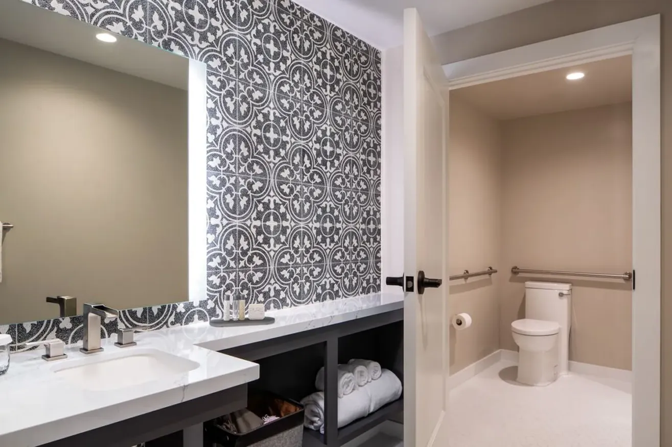 Modern bathroom with a patterned black and white accent wall, large illuminated mirror, marble countertop, open shelves with towels, and a separate toilet area with grab bars and beige walls.