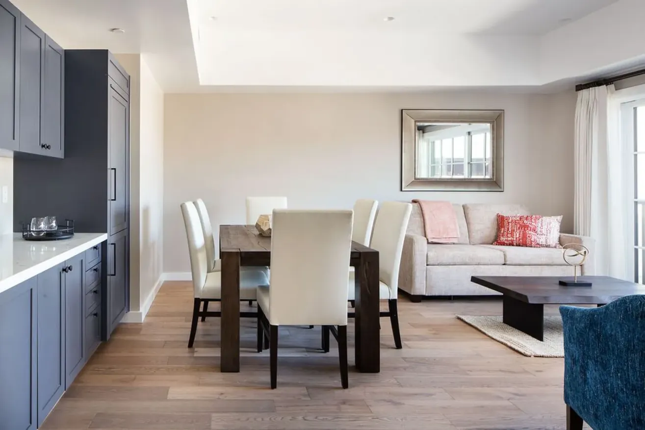 Modern open-plan living and dining area with gray cabinets, a dark wood dining table, cream chairs, a beige sofa with red cushions, a coffee table, and large windows letting in natural light.