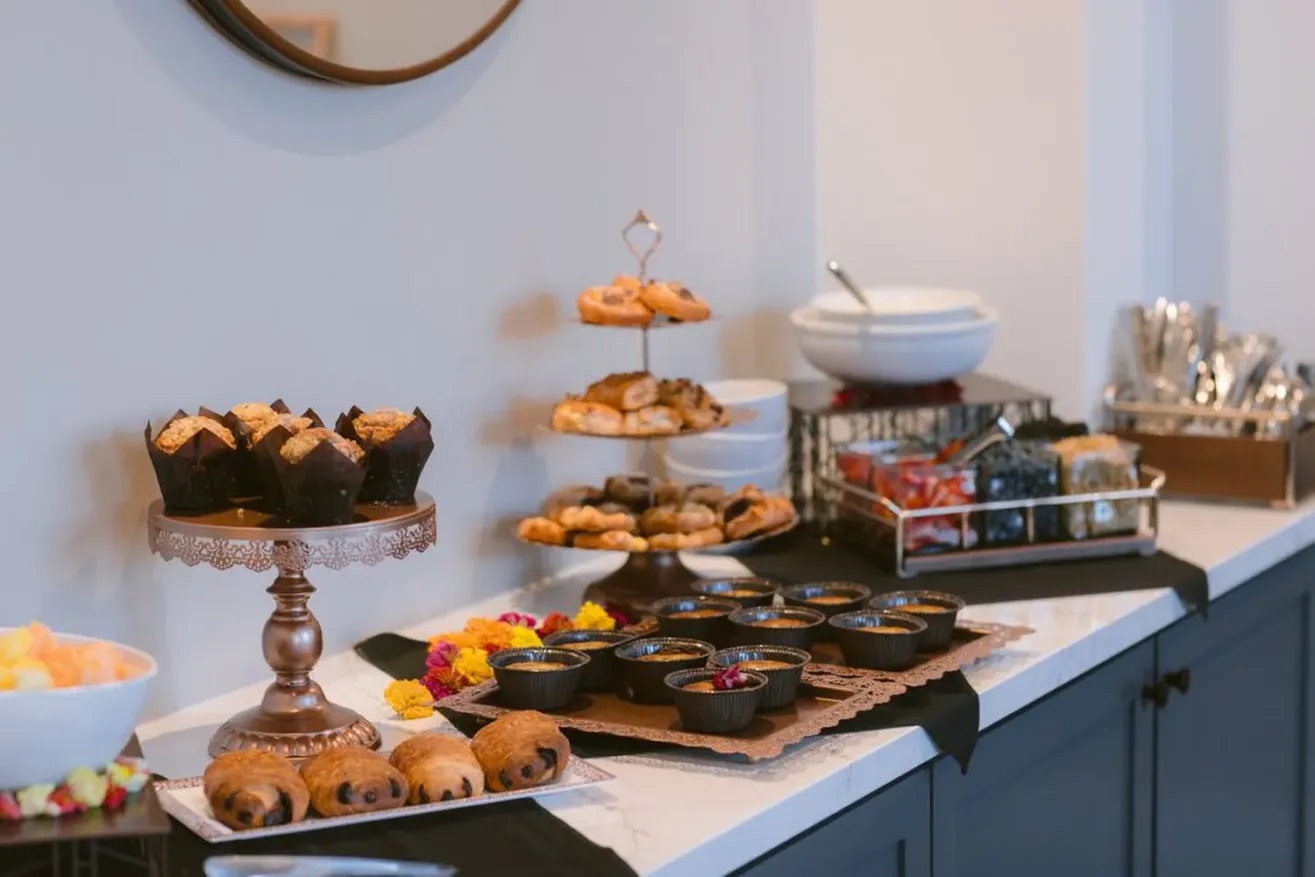 A buffet table at The Piccolo at Paso Robles Inn features assorted pastries, muffins, fruit bowls, and desserts on elegant trays and stands, with stacked plates, napkins, and cutlery set neatly to the side against a white wall.