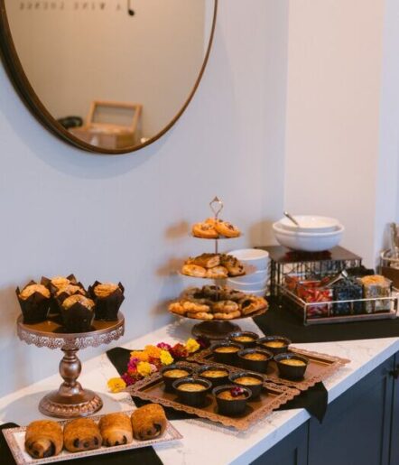 A countertop at The Piccolo at Paso Robles Inn displays a variety of pastries, including muffins, croissants, and tarts on trays and stands. A three-tiered stand holds cookies, with colorful flowers as decoration and a mirror hanging above.