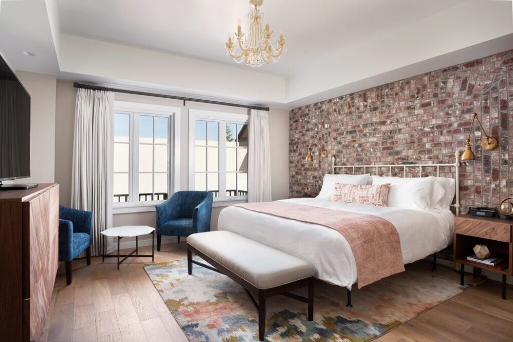 A stylish bedroom, inspired by suites in Paso Robles, features a large bed, brick accent wall, blue chairs by the window, a light rug over wooden floors, modern side tables, a chandelier, and a cushioned bench. Natural light fills the room.