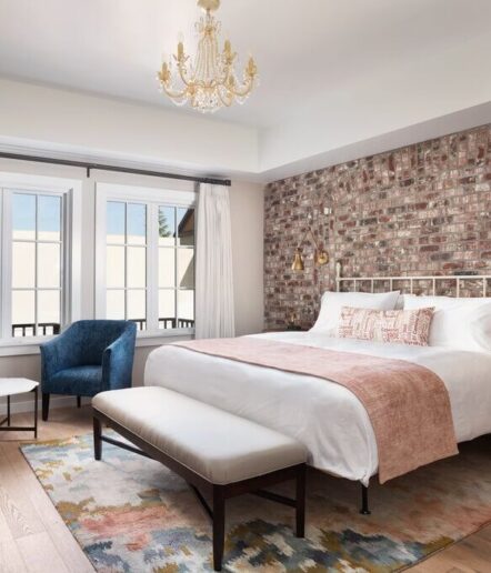 A cozy bedroom reminiscent of suites in Paso Robles, with a brick accent wall, white bed and pink blanket, blue armchair, colorful rug, and gold chandelier. Large windows bathe the space in natural light through partially drawn curtains.