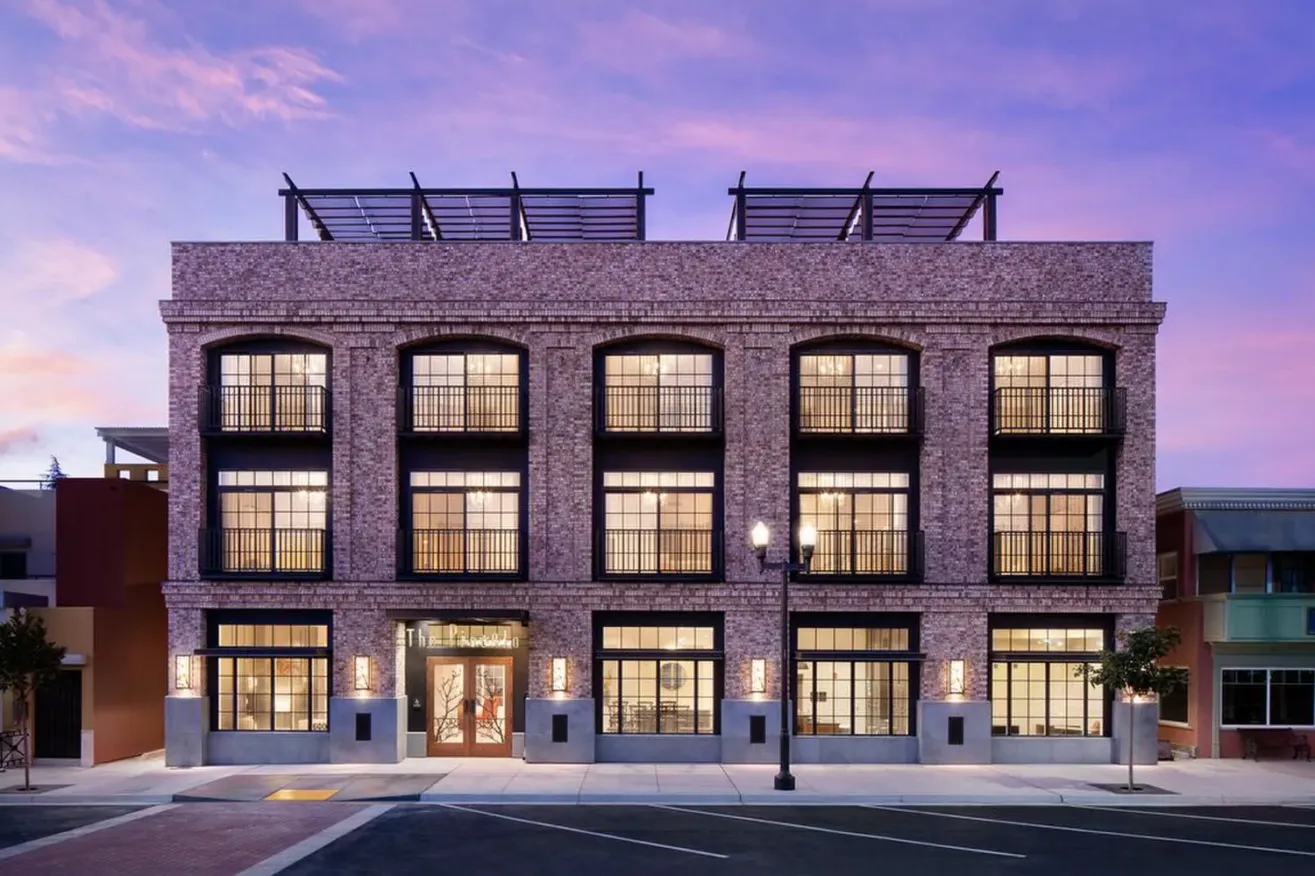 A three-story brick building with large, evenly spaced windows and black railings, illuminated from within, stands on a quiet street under a sunset sky with purple and pink hues—offering elegant suites in Paso Robles.