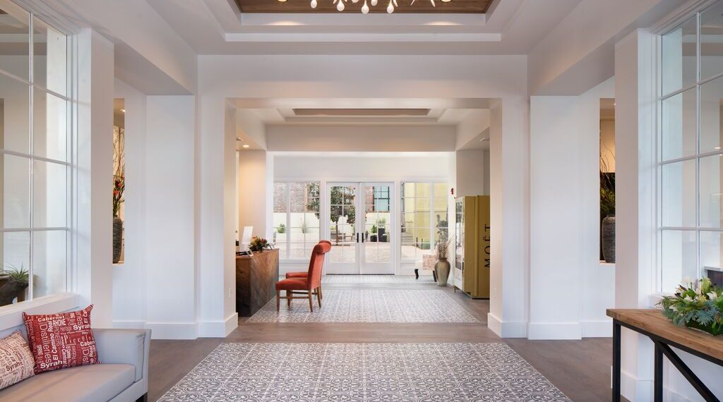 A bright, modern lobby at The Piccolo at Paso Robles Inn features patterned tile flooring, white walls, large windows, a red chair at a wooden desk, and decorative plants leading to glass doors that open to an outdoor area.