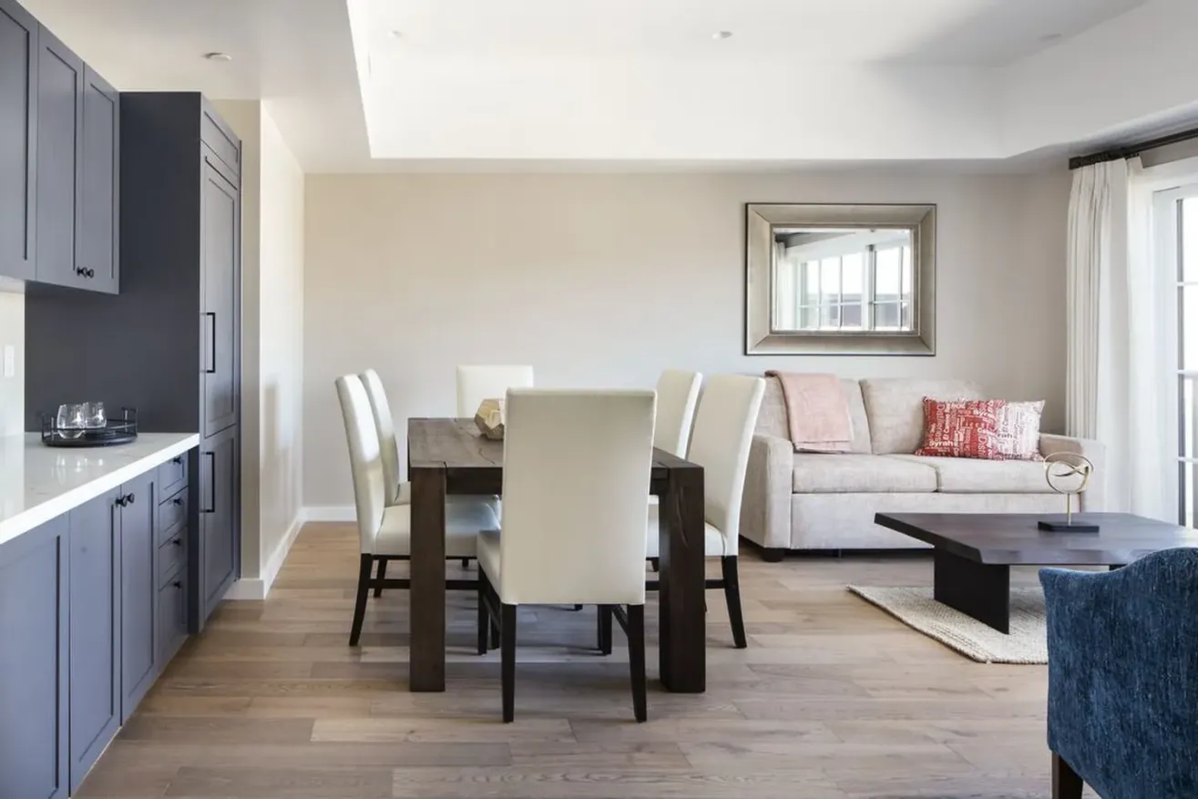 Modern open-plan living and dining area with light wood floors, a dark dining table surrounded by cream chairs, a beige sofa with red and pink cushions, a blue armchair, and a large wall mirror.