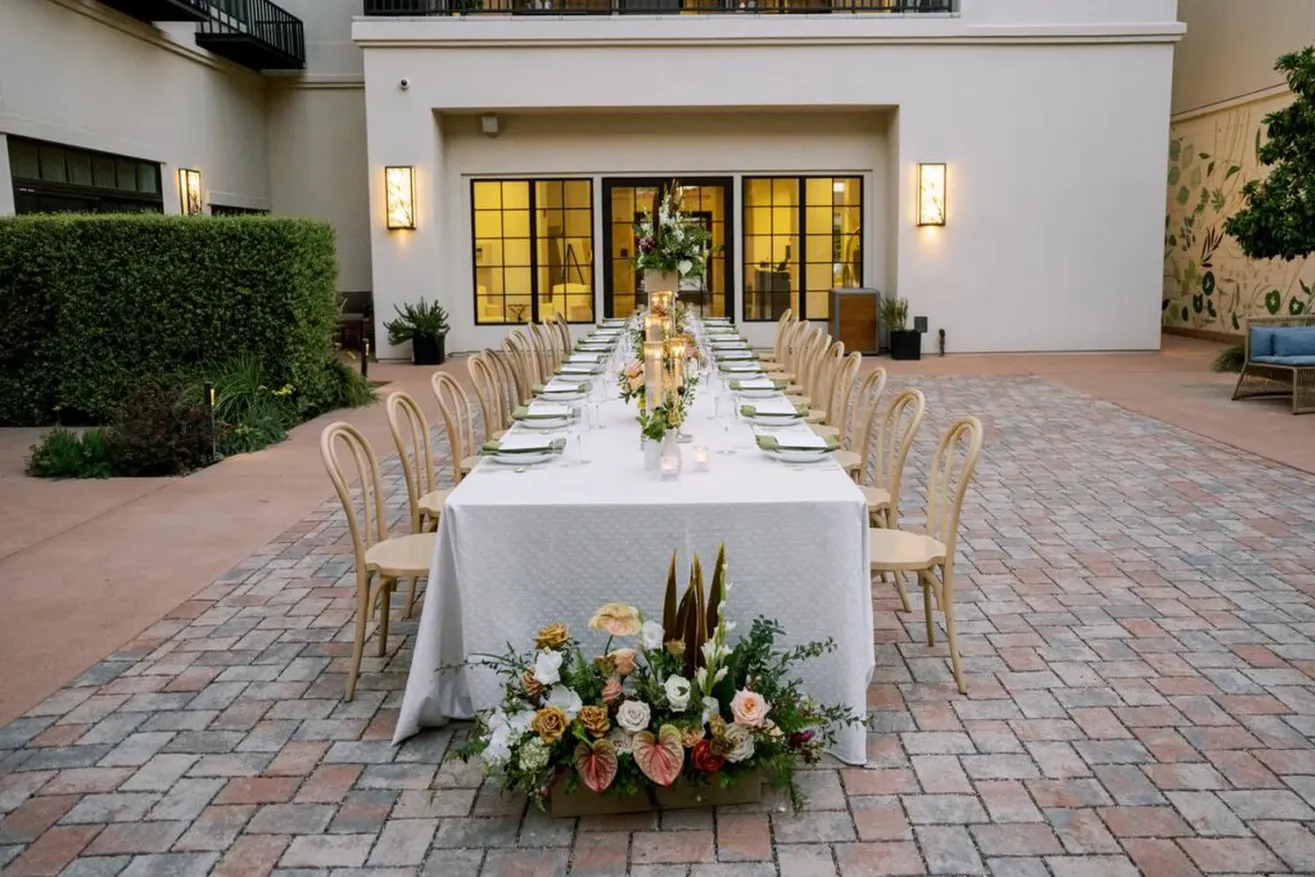 A long, rectangular outdoor dining table with a white tablecloth is set for a formal event at The Piccolo at Paso Robles Inn. Wooden chairs line both sides, floral centerpieces adorn the table, and a modern building frames the courtyard setting.