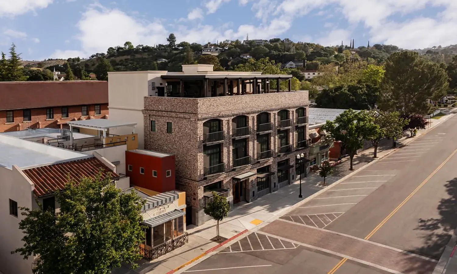 A modern brick building with balconies and rooftop seating stands on a quiet street lined with trees, with hills and greenery in the background under a partly cloudy sky.