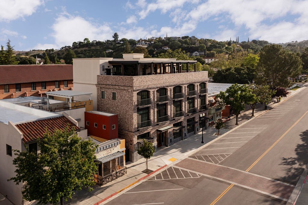 A modern brick building with balconies and rooftop seating stands on a quiet street lined with trees, with hills and greenery in the background under a partly cloudy sky.