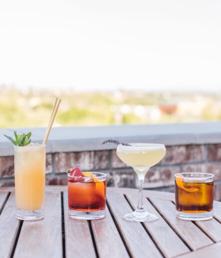 Four colorful cocktails in different glasses sit on a wooden outdoor table, with a blurred scenic background. Each drink is garnished uniquely with mint, berries, citrus, or a lavender sprig.