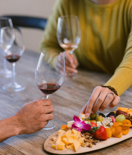 Two people sit at a wooden table at The Piccolo at Paso Robles Inn, each holding a glass of red wine. One reaches for cheese from a platter with fruits, nuts, and cheese slices, all beautifully garnished with an edible flower.