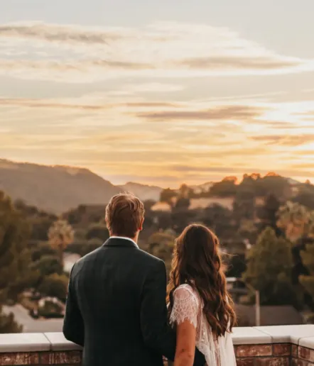 A couple stands close together, facing away and looking at a scenic sunset over rolling hills and trees, with soft clouds in the sky.