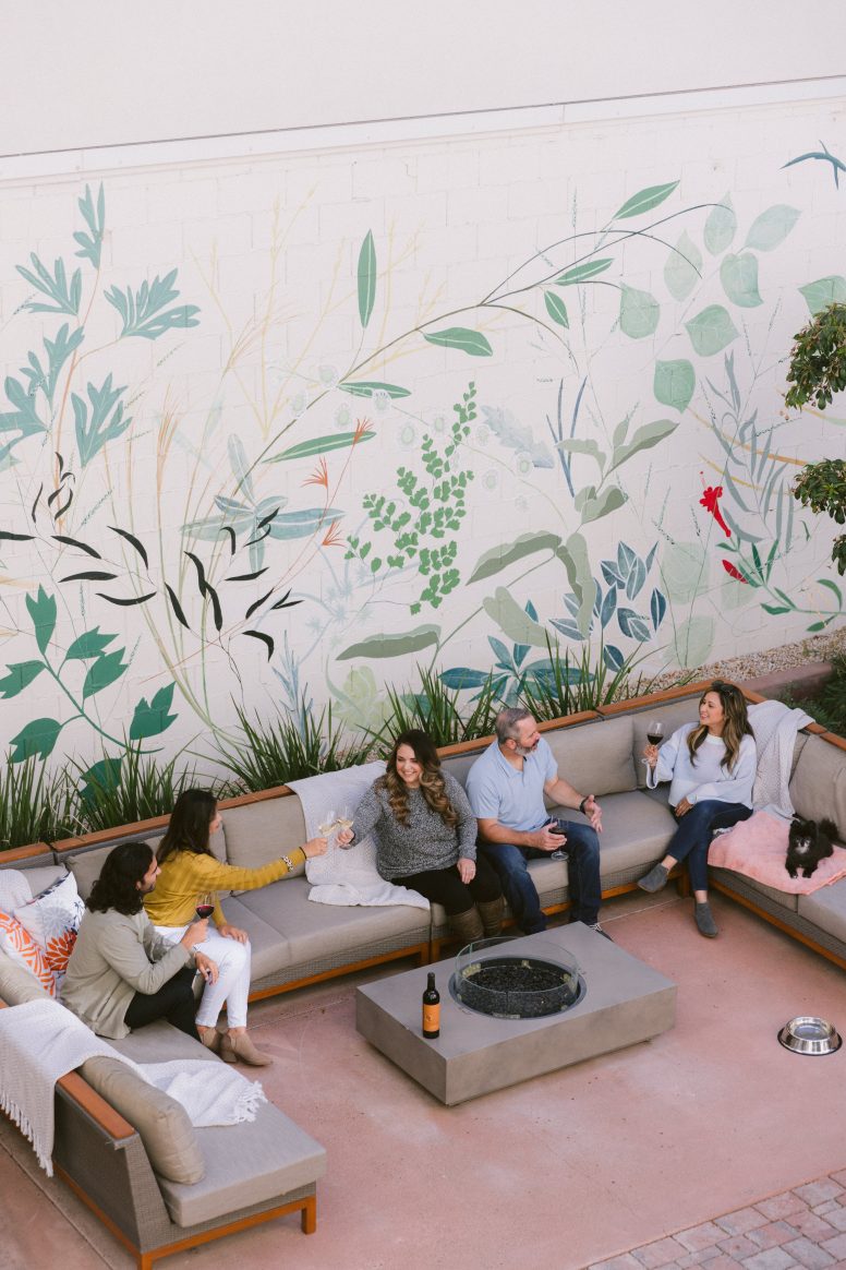 Four people sit and chat on outdoor patio furniture around a fire pit, with a dog nearby. A large mural of plants and leaves decorates the wall behind them, creating a lively and cozy atmosphere.