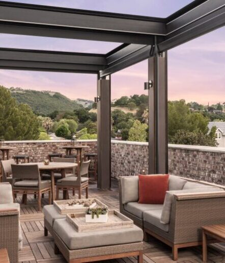 Outdoor patio with wicker furniture, cushioned chairs, and tables on a wooden deck. Surrounded by a brick half-wall, the area overlooks green hills under a pastel sunset sky.