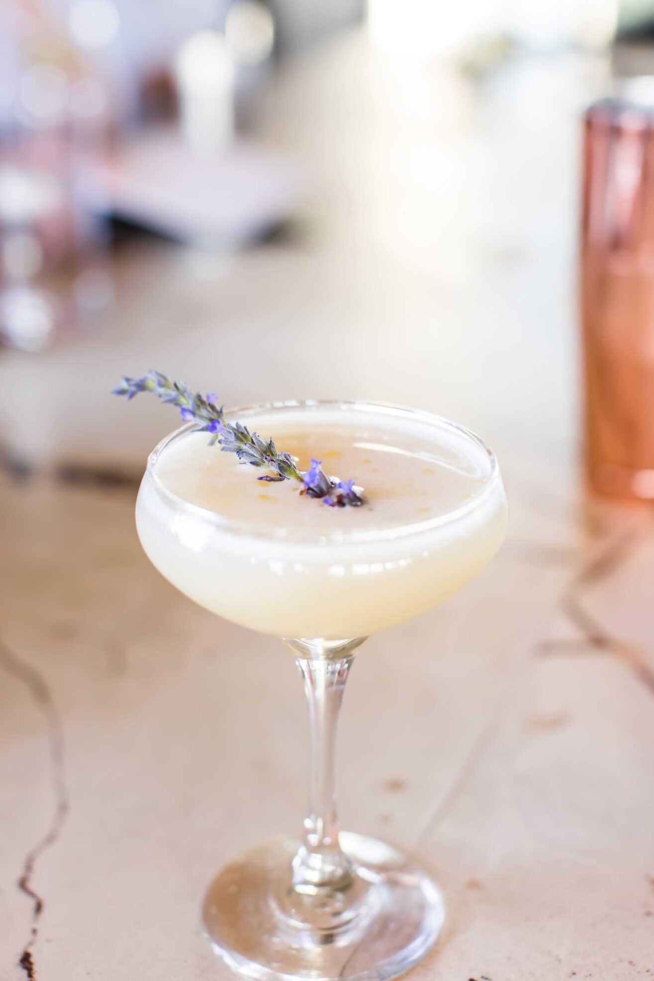 A coupe glass filled with a pale yellow cocktail, garnished with a small sprig of lavender flowers, sits on a light marble surface. The background is softly blurred.