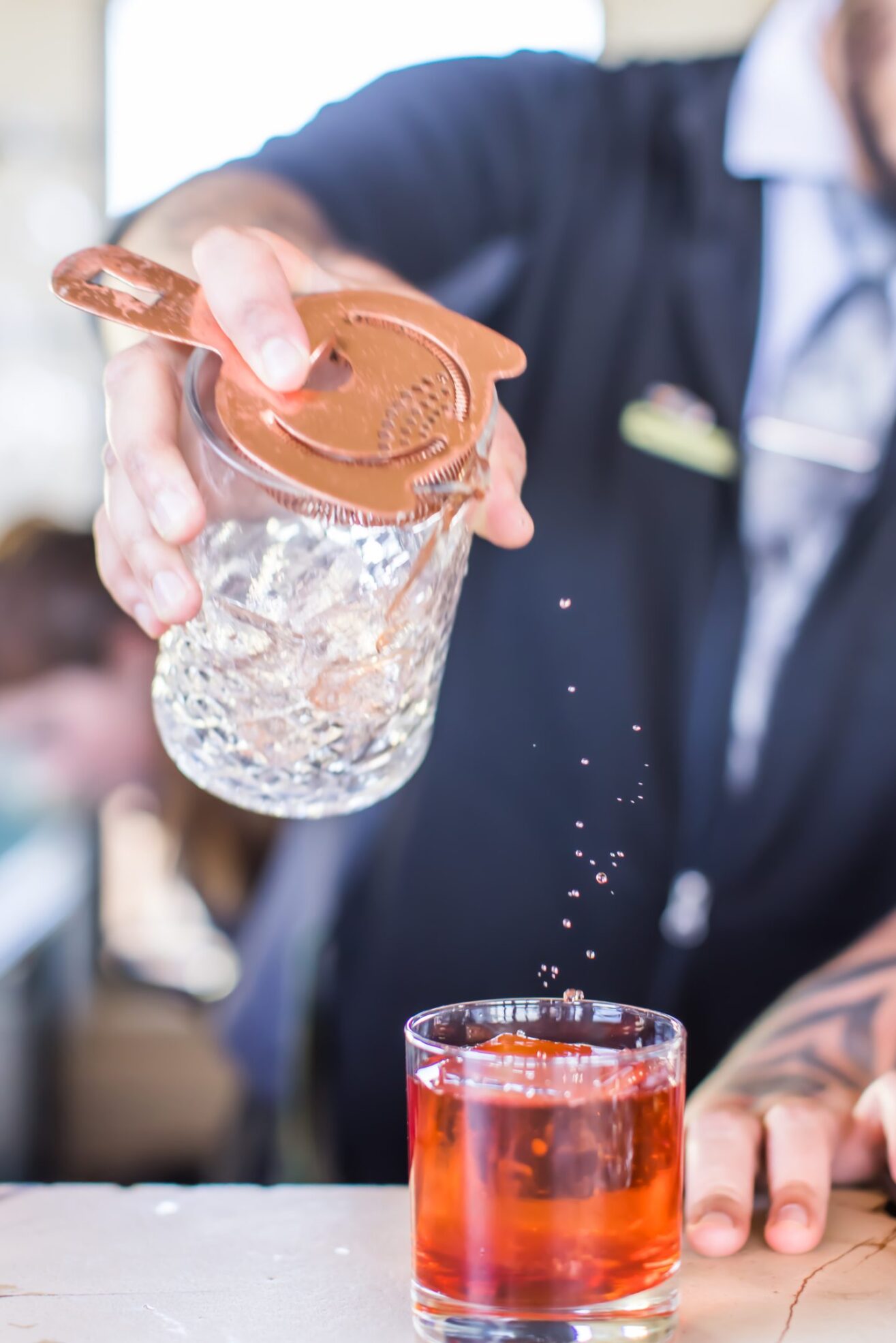 A bartender pours a cocktail from a mixing glass with a strainer into a glass filled with red liquid, with droplets splashing above the drink. The bartenders hand and part of their uniform are visible.