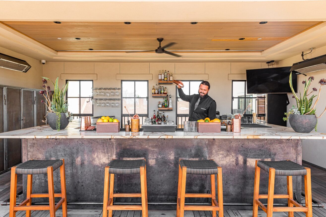 A bartender stands behind a rooftop bar, preparing a drink. The bar has four empty stools, potted plants, citrus fruits, and bottles. Large windows and a TV are in the background, with bright lighting.
