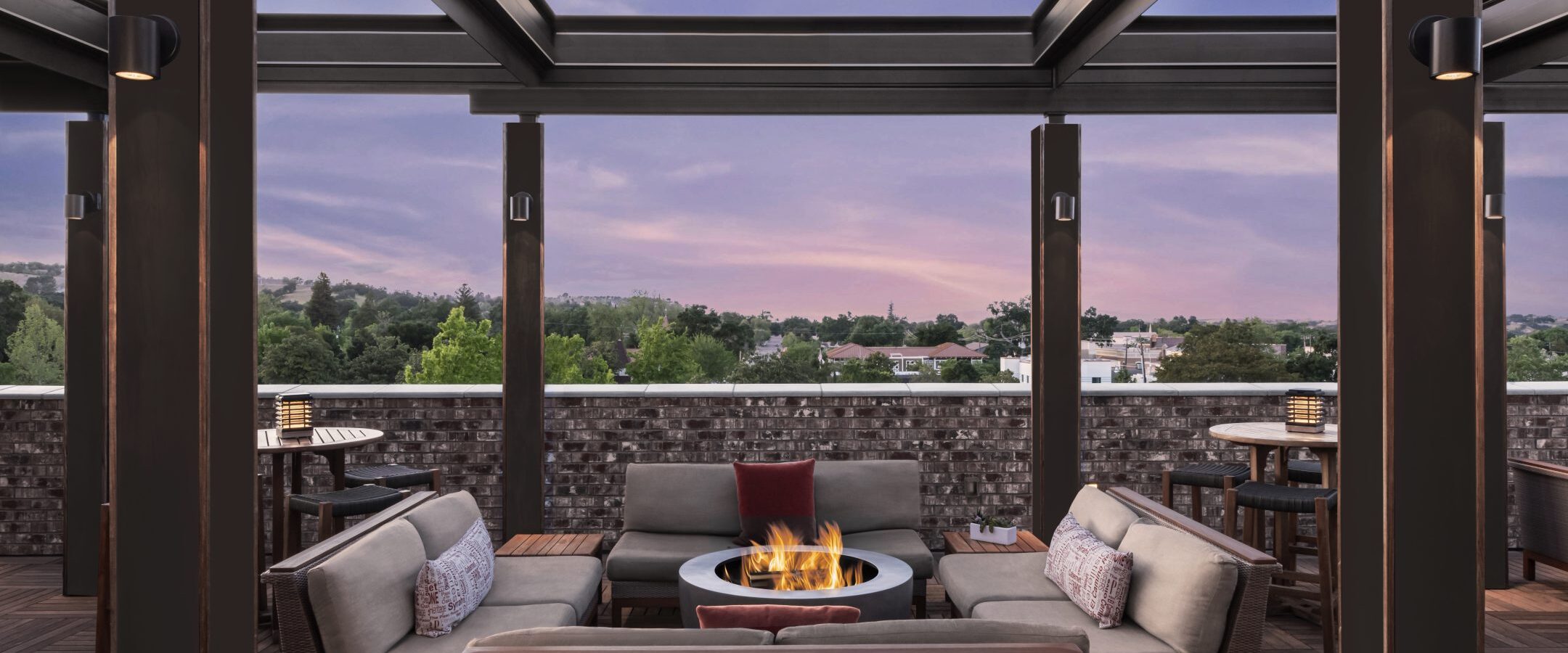 Rooftop lounge at sunset with modern sofas around a fire pit, under a pergola. Trees and buildings are visible in the distance, creating a relaxing outdoor atmosphere.