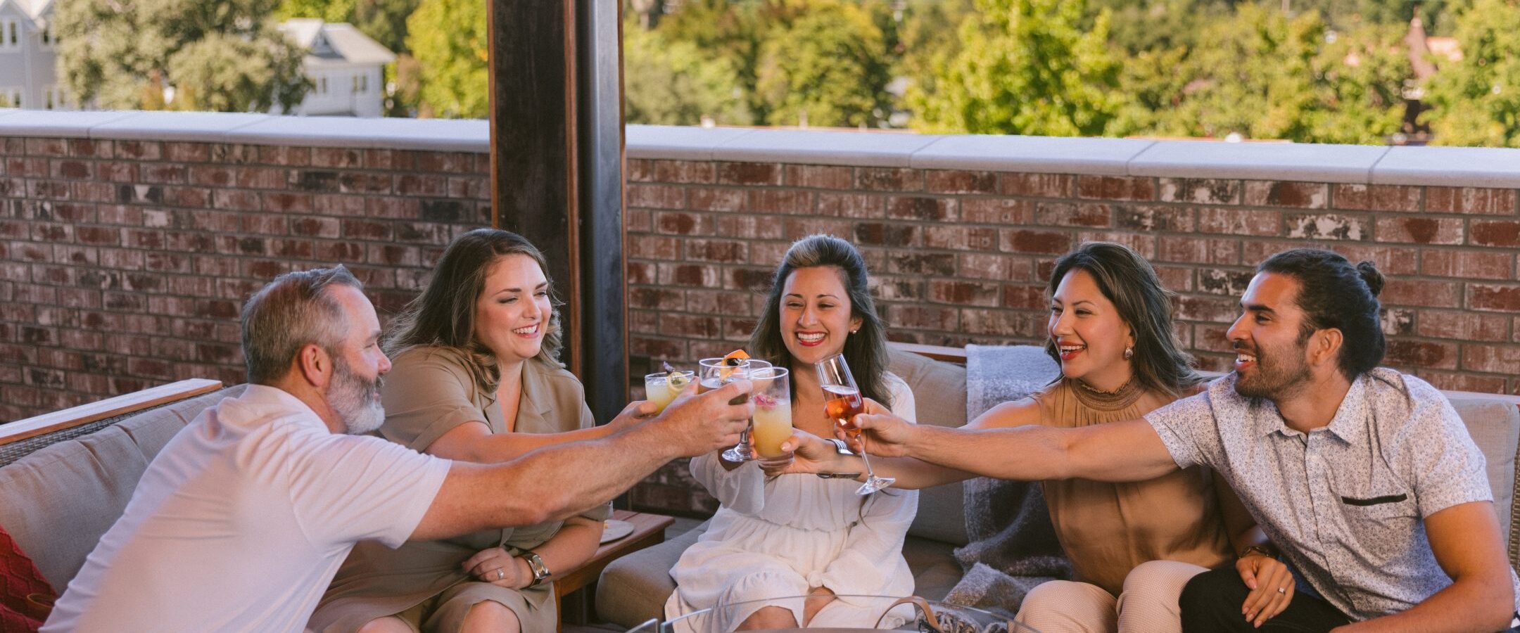 Five people sitting on an outdoor patio sofa, smiling and clinking drinks together around a fire pit table, with trees and houses in the background on a sunny day.
