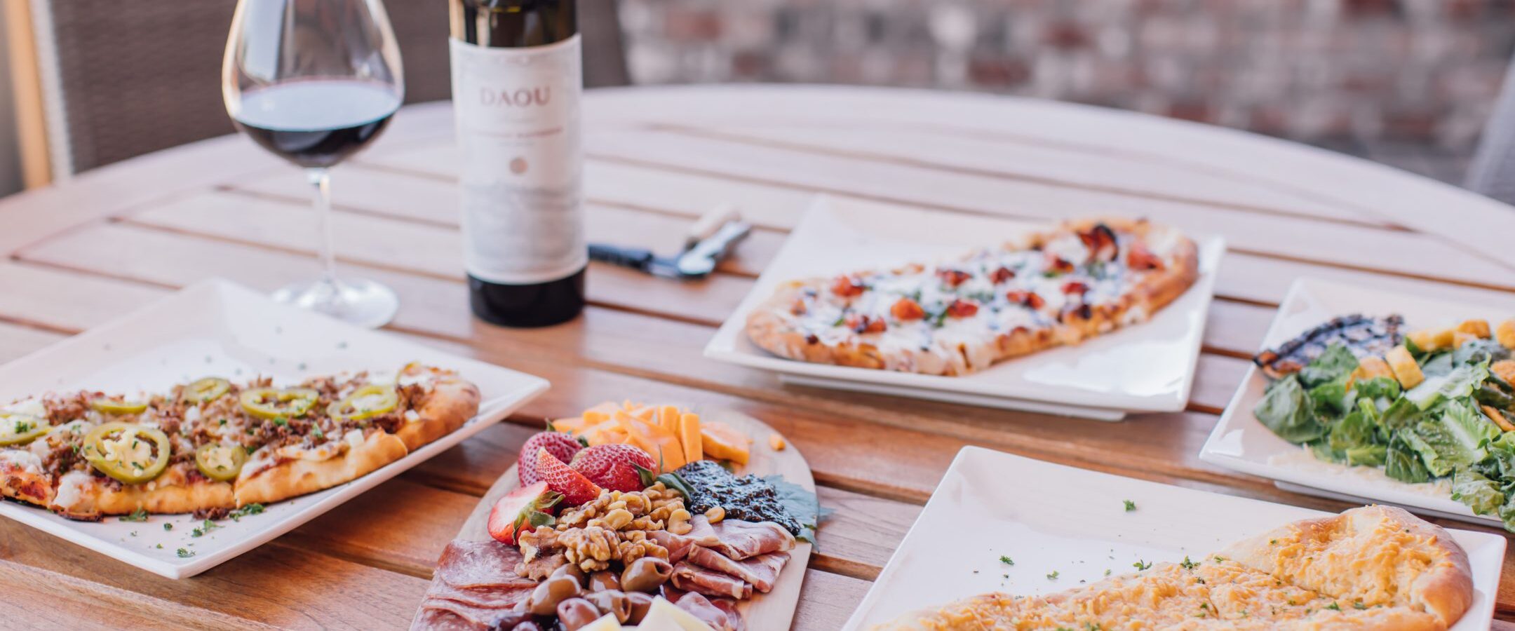 A round wooden table set with flatbreads, a charcuterie board with cheese, cured meats, nuts, and fruits, a plate of salad, a bottle of red wine, and a filled wine glass. Brick wall background.