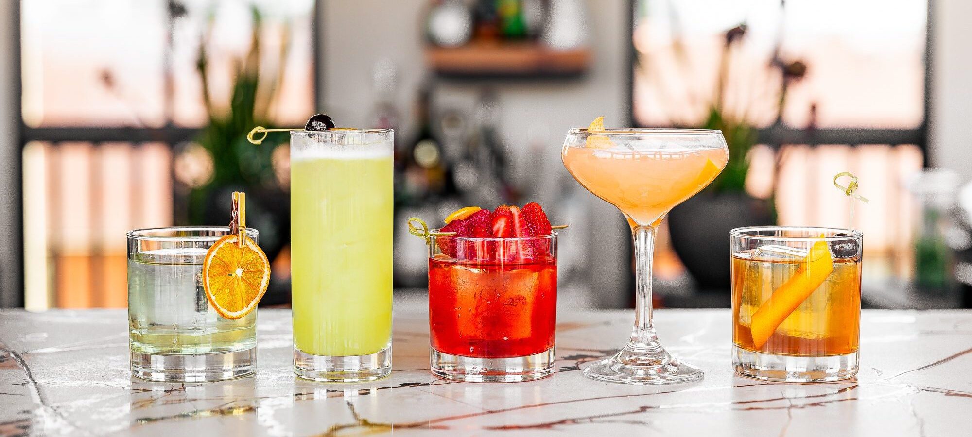 Five colorful cocktails in assorted glassware sit on a marble bar, each garnished with fruit slices or berries, with a blurred background showing windows and plants.