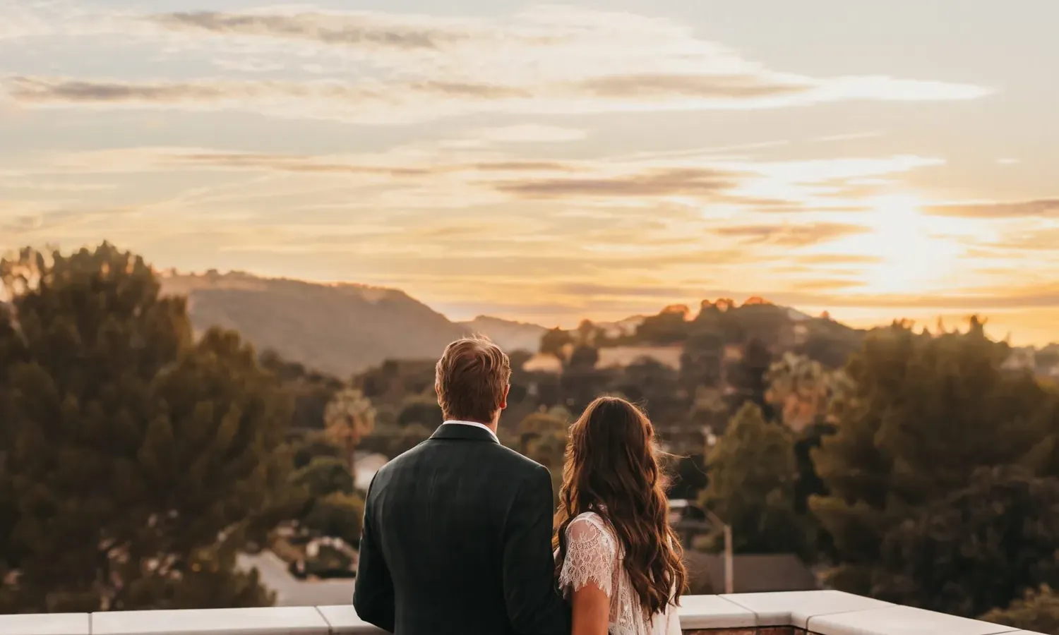 A couple stands side by side on a balcony, looking at a scenic sunset over rolling hills and trees. The sky is partly cloudy with warm, golden light illuminating the landscape.