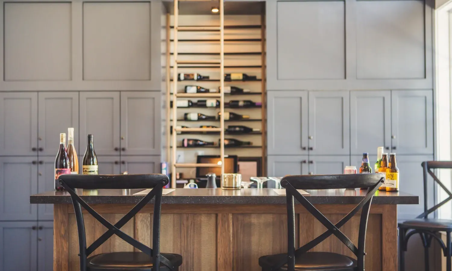 A modern home bar with a wooden island, two black chairs, several bottles of alcohol on the counter, and built-in shelves behind showcasing bottles and gray cabinets.