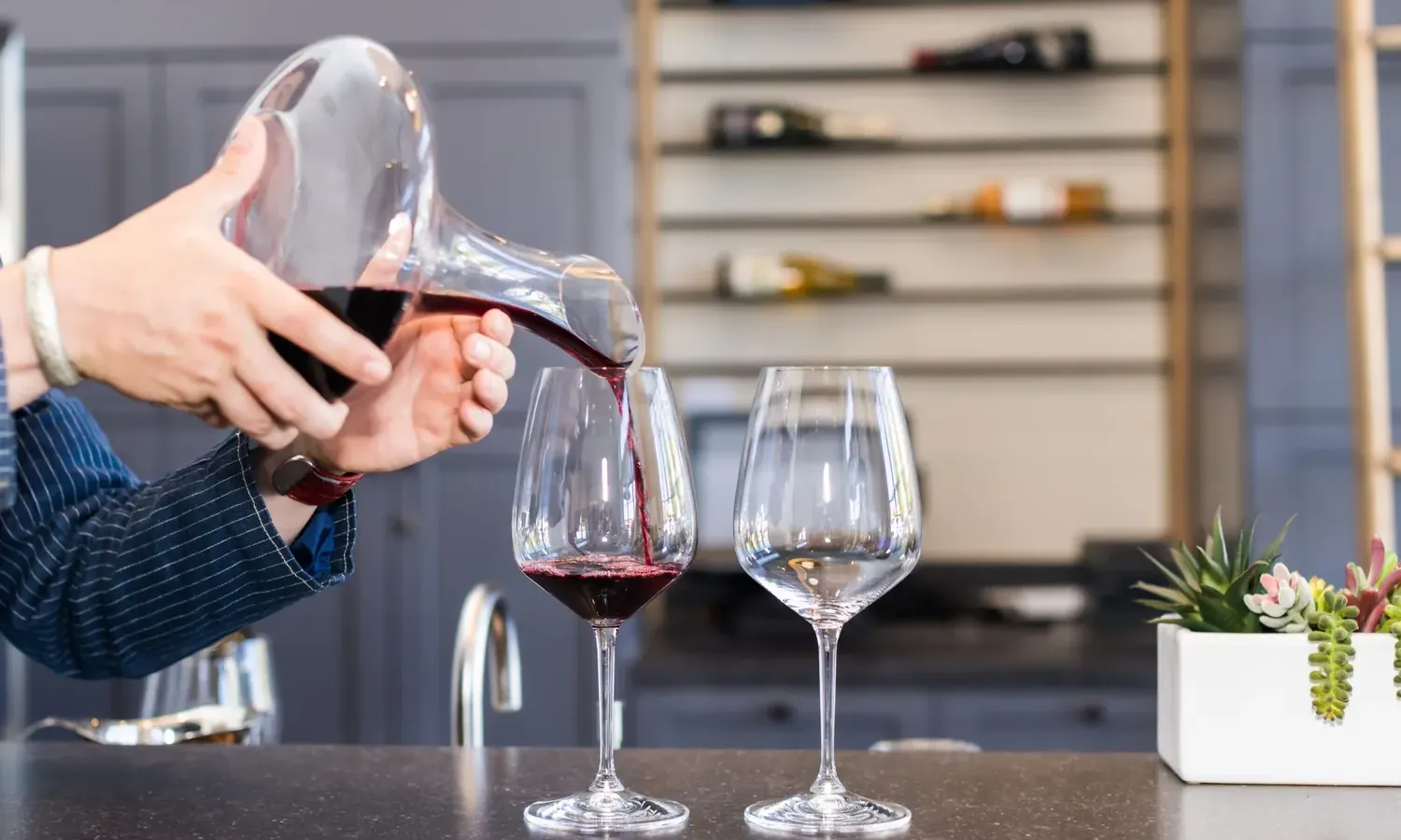 A person pours red wine from a decanter into a wine glass on a counter, with another empty glass beside it. Shelves with wine bottles and a small potted plant are in the background.