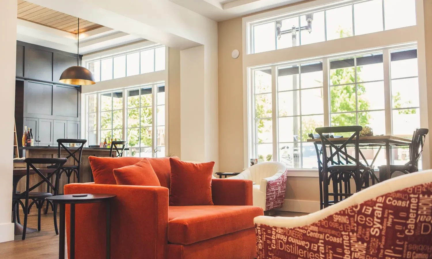 A modern, sunlit living room with large windows, an orange armchair, patterned chairs, wooden floors, and a dining area in the background with dark furniture and a hanging light fixture.