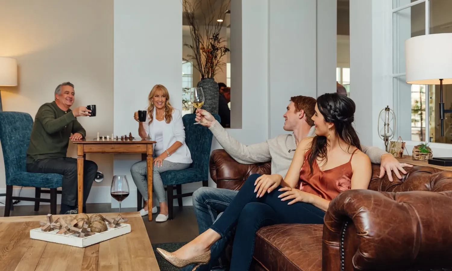 Four adults relax in a cozy living room; two sit on a leather couch toasting with white wine, while two others sit at a chess table, smiling and holding mugs. The room is warmly lit and inviting.