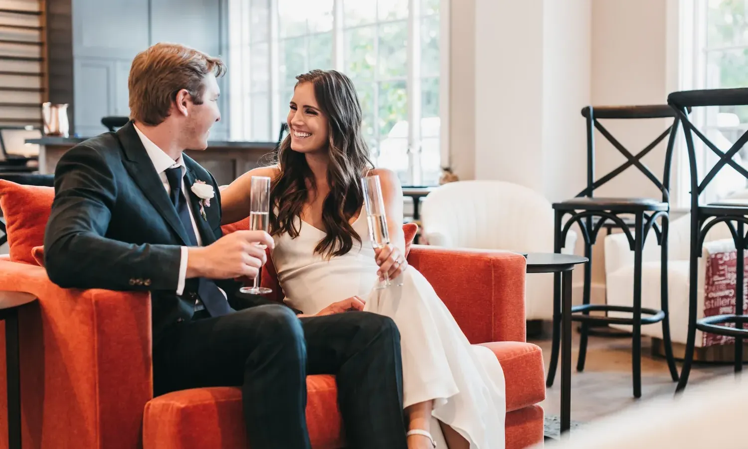 A smiling couple in formal attire sit close together on a red couch, holding champagne glasses and looking at each other warmly in a bright, modern room with large windows.