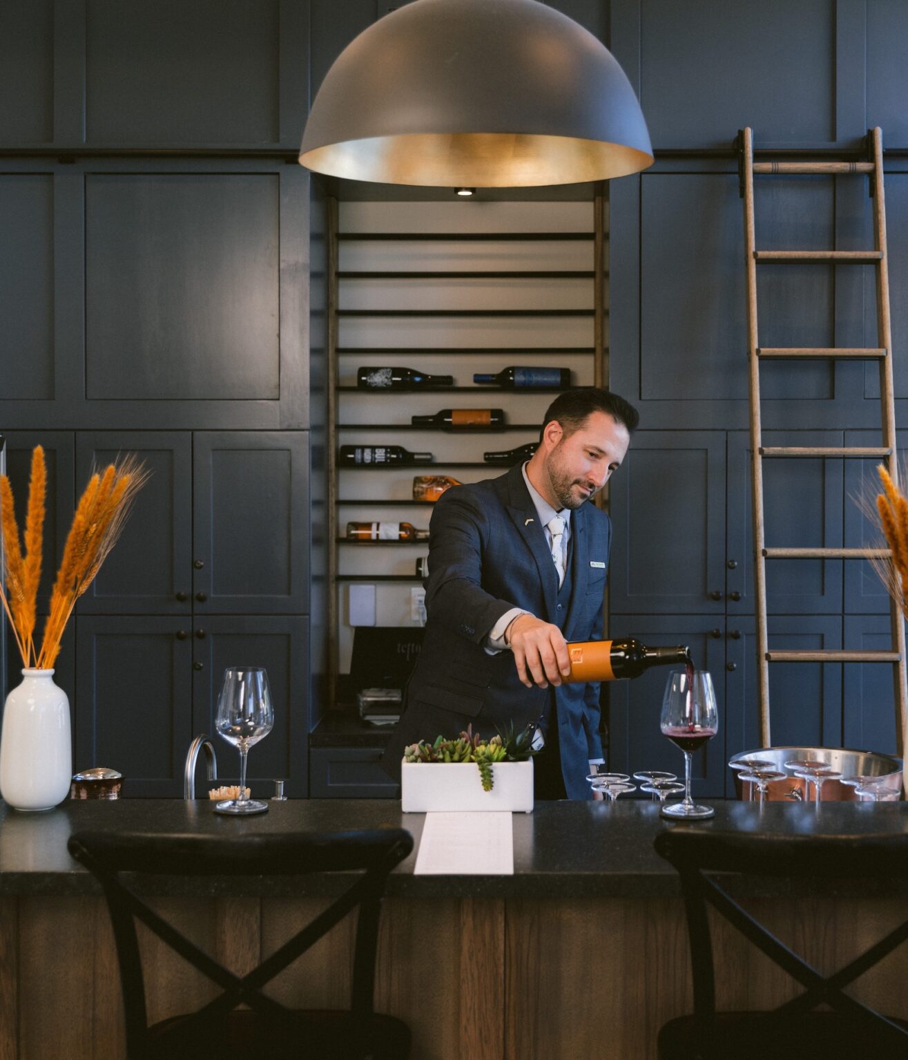 A man in a suit pours red wine into a glass at a modern wine bar with dark cabinets, a large dome pendant light, a ladder, and decorative vases with dried grasses. Bottles of wine are displayed on shelves behind him.