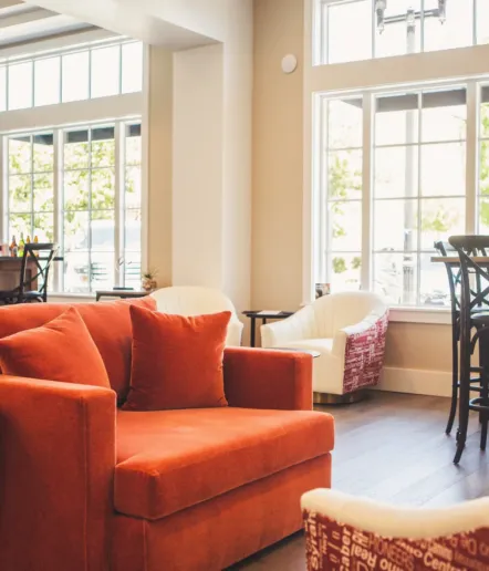A cozy, modern living room with a large orange armchair, white accent chairs, and tall windows letting in natural light. The space features wooden floors and a welcoming, comfortable atmosphere.
