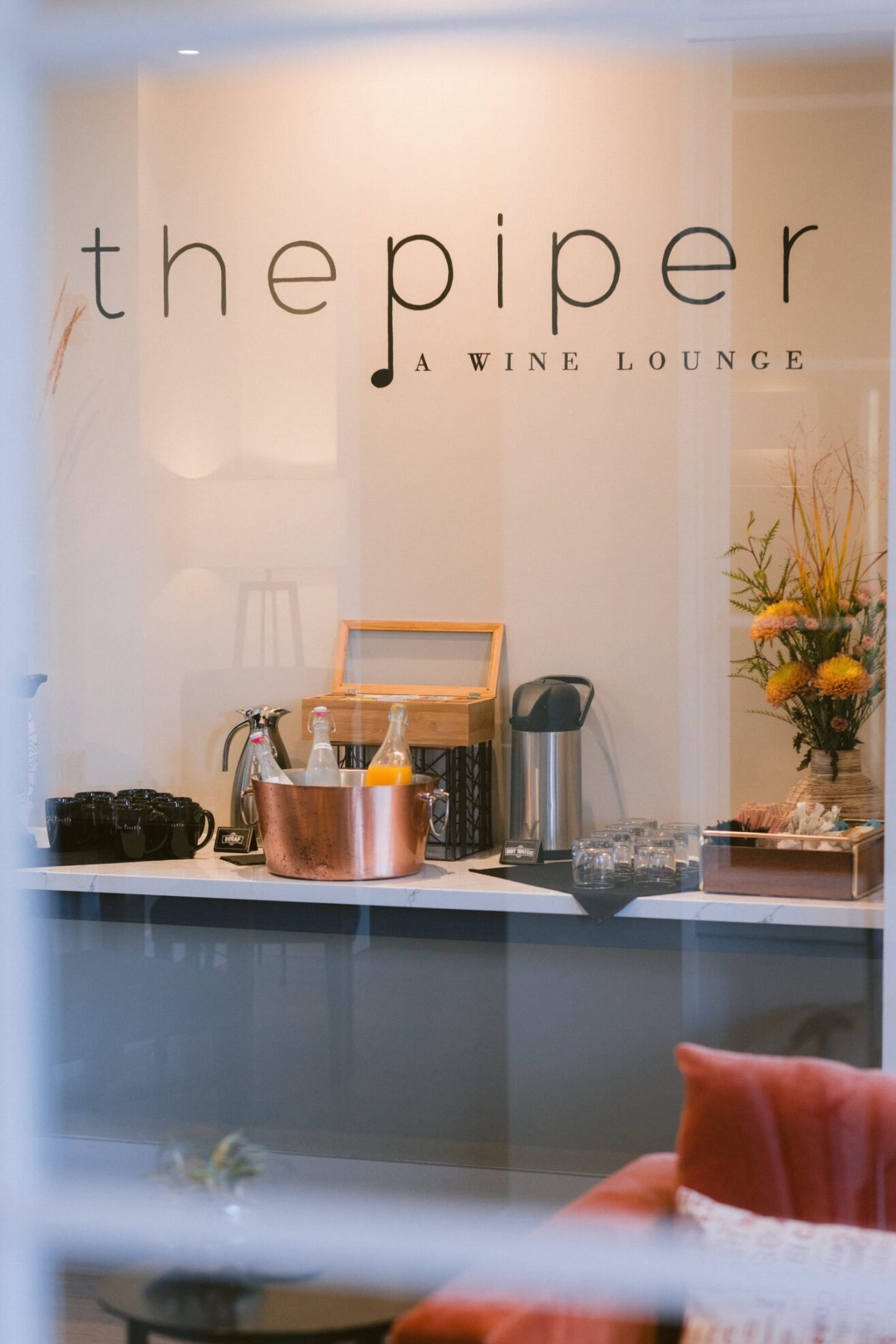 View through a window of The Piper: A Wine Lounge with a counter displaying a copper ice bucket with drinks, a coffee pot, glassware, and a flower arrangement next to a cozy seating area.